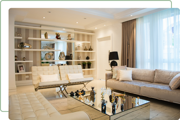 fancy living room with multiple plush sofas, a mirror coffee table and a wall of shelves with decore, and a large window in the back covered with white mesh curtains